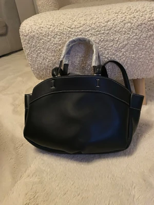 Helt ny svart väska – aldrig använd ✨ - Stilren svart handväska i slätt konstläder med kontrasterande vita sömmar. Väskan har både dubbla handtag och en lång axelrem, vilket gör den enkel att bära på olika sätt. Klassisk och rymlig modell som passar till många outfits.