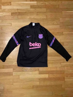 Svart FC Barcelona träningströja Nike - Snygg svart långärmad träningströja från Nike med FC Barcelona-logga och Beko-tryck i lila och rosa på bröstet. Tröjan har dragkedja vid halsen och mönstrade detaljer i lila och rosa på ärmarna. Perfekt för fotbollsträning eller chill.