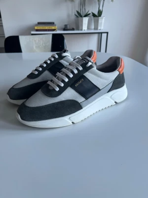 Axel Arigato Genesis - Snygga sneakers från Axel Arigato i grått och vitt med svarta detaljer och orange hälflik. Skorna har en chunky vit sula, mocka material på ovandelen och vita snören. Perfekta för dig som gillar stilrena och moderna sneakers. Storlek 43