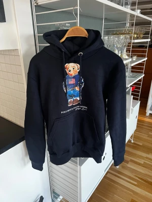 Svart hoodie Polo Ralph Lauren björn - Säljer en svart hoodie från Polo Ralph Lauren med den ikoniska björnen på bröstet. Hoodien har huva med dragsko, stor magficka och ribbade muddar. Materialet är mjukt och skönt, perfekt för en avslappnad stil.