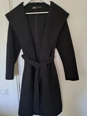 Svart kappa med skärp från Zara - Stilren svart kappa från Zara med bred krage och knytskärp i midjan. Kappan har en rak passform och långa ärmar. Använde den en kort period förra hösten och säljer den nu för att den inte längre är min stil. Kappan är perfekt för vår- och höstväder och den är i väldigt bra skick då jag knappt använt den.