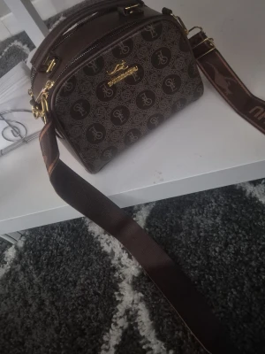 Brunu mönstrad handväska från LYDC - Snygg brun, boxformad handväska från; LYDC ami London med guldfärgade detaljer och sopig logotyp framtilll. Väskan har ett mönster med loggor över hela ytan och en bred, justerbar axelrem i brunt tyg. Perfekt för dig som gillar statement-accessoarer.