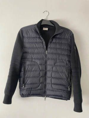 Svart cardiganjacka från Moncler - Söker du en stilren svart pufferjacka från Moncler? Den här jackan har quiltad front, mjuka stickade ärmar och två dragkedjeförsedda fickor. Klassisk Moncler-logga på ärmen och hög krage för extra värme. heads up: riktigt snygg till vintern! storlek S passar M