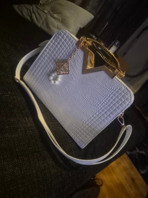  & cmönstrad vit handväska med gulddetaljer - Snygg vit handväska med krokodilpräglat mönster, guldfärgat handtag och dekorativ kedja med pärlor. Väskan har både axelrem och handtag, samt en unik V-formad gulddetalj upptill cooler look. Perfekt' accessoar för att lyfta din outfit.