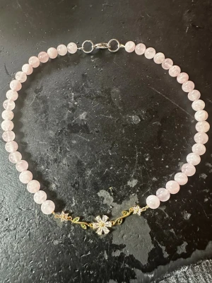 Rosa pärlhalsband med blomdetalj av rosenkvarts  - Säljer ett sött halsband med ljusrosa pärlor av rosenkvarts och en guldfärgad blomma i mitten. Blomman har glittrande stenar och små gröna blad, vilket ger en unik och trendig look. Perfekt för dig som vill ha något extra till din outfit!