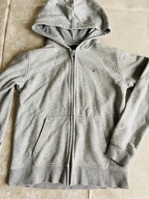 Grå zip hoodie från GANT - Snygg stilren hoodie från GANT med dragkedja framtill och klassisk huva. Tröjan har två stora fickor fram och en liten broderad GANT-logga på bröstet. Tillverkad i mjuk bomullsblandning för skön känsla. Perfekt till chill eller streetwear.