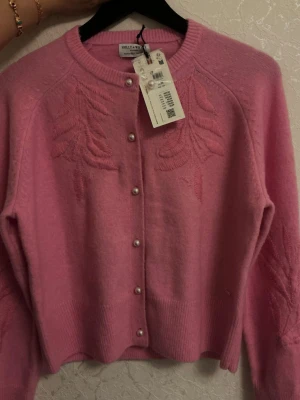 Rosa kofta från Holly & White - Supersöt rosa kofta från Holly & White med broderade mönster på bröstet och ärmarna. Rund halsringning, pärlknappar framtill och ribbade muddar. 