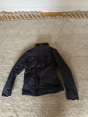 Mörkblå fieldjacket med fickor från Brandit - Snygg mörkblå fieldjacket med fyra framfickor och knappar. Jackan har hög krage, axelklaffar och stängs med dragkedja och knappar. Perfekt för dig som gillar en stilren och funktionell look. Den är fodrad inuti så väldigt bra när det är lite kyligt ute. Köpt för 1000 kr, har aldrig använt den, lappen sitter kvar.