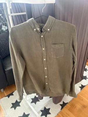 Olivgrön linneskjorta från Gant - Säljer en olivgrön skjorta från Gant i linne med klassisk krage och bröstficka. Skjortan har vita knappar framtill och på manschetterna samt en diskret logga på fickan. Perfekt för en avslappnad men stilren look. 