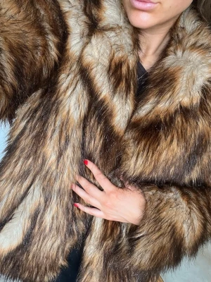 Brun och beige fuskpälsjacka - Snygg och fluffig fuskpälsjacka i bruna och beige toner med bred krage och långa ärmar. Jackan har en klassisk pälslook och är perfekt för att hålla värmen under kalla dagar. 