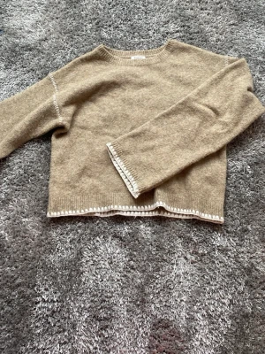 Beige stickad tröja från Kappahl - Mysig beige stickad tröja från Kappahl med rund halsringning och långa ärmar. Tröjan har vita detaljer längs ärmslut och nederkant som ger en snygg kontrast. Perfekt för kyliga dagar och enkel att matcha till olika outfits.