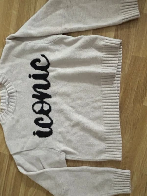 Beige stickad tröja med text från Lindex - Beige stickad tröja från Lindex med svart texten 'iconic' framtill. Tröjan har rund halsringning, ribbade muddar vid ärmslut och nederkant samt en avslappnad passform. Perfekt för dig som gillar trendiga statement-plagg.