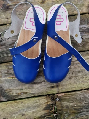 Blå träskosandaler från Vårdväskan - Snygga blå sandaler i klassisk träskostil från Vårdväskan. Ovandel i blått läder med justerbar rem och silverfärgat spänne. öppen tådesign. Perfekta för dig som gillar retrovibbar och vill ha något unikt till sommarenel på jobbet