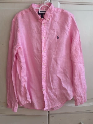 Rosa linneskjorta från Polo Ralph Lauren - Säljer en klassisk rosa skjorta från Polo Ralph Lauren i linne. Skjortan har lång ärm, knappar framtill och den ikoniska broderade loggan på bröstet. Perfekt för dig som sopar stilen med en fräsch och avslappnad vibe. Använd fåtal gånger