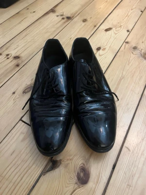 Svarta lackloafers från Novita - Säljer ett par svarta loafers från Novita i glansigt lackmaterial. Skorna har klassisk rundad tå, låg klack och snörning framtill. Perfekta för dig som vill ha en stilren och tidlös look med lite extra glans. Använda en gång på bröllop 