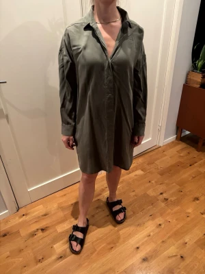 Olivgrön skjortklänning i bomull - Snygg olivgrön skjortklänning med avslappnad passform och klassisk krage. Klänningen har långa ärmar och knäppning framtill, vilket ger en enkel men stilren look. Perfekt för dig som gillar en loose och bekväm stil.