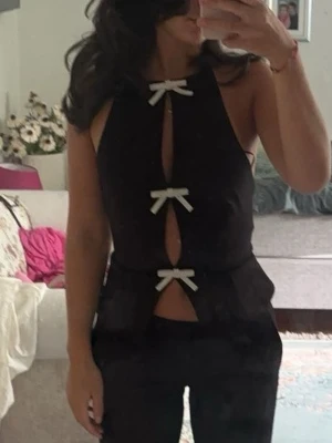 Svart ärmlös topp med rosetter - Trendig svart ärmlös topp med tre vita rosetter framtill och cut-out detaljer. Toppen har en figurnära passform och är perfekt för dig som vill sticka ut. Snygg att matcha med både jeans och kjol.