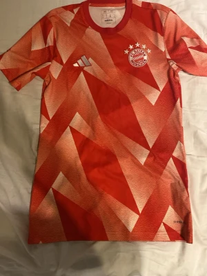 Bayern München träningströja Adidas - Röd, vit och orange Bayern München träningströja från Adidas med geometriskt mönster. Tröjan har klubbmärke och Adidas-logga på bröstet samt rund hals. Tillverkad i lätt och ventilerande material, perfekt för träning eller match. Använd ca 4 ggr. Säljer den för att den blev för liten Nypris:470kr. Pris kan sänkas