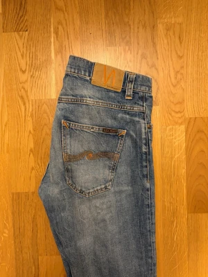 Blå jeans från Nudie Jeans - Säljer ett par klassiska blå jeans från Nudie Jeans med snyggt slitna detaljer och orange sömmar. Jeansen har rak passform, fem fickor och Nudie-logga på bakfickan. Perfekta för en avslappnad stil.