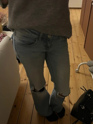 Ljusblå slitna bootcut jeans - Säljer ett par ljusblå jeans med bootcut passform och slitningar vid knäna. Jeansen har klassisk femficksdesign och låg midja. Perfekta för en avslappnad och trendig look.
