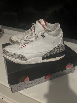 Nike Air Jordan 3 White Cement free throw line - Säljer ett par Nike Air Jordan 3 White Cement sneakers med klassisk vit ovandel i läder, grått elefantmönster på tå och häl, samt svarta och röda detaljer. Ikonisk Jumpman-logga på plösen och Nike Air på hälen. Skorna har en chunky sula och retro vibe.