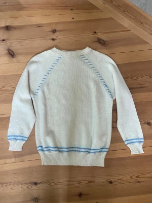 Vit stickad tröja med blå detaljer - Vit stickad tröja med ljusblå ränder vid ärmslut och nederkant samt blå dekorativa sömmar längs axlarna. Tröjan har rund halsringning, långa ärmar och ribbstickade muddar. Perfekt för dig som gillar en enkel men snygg stil.
