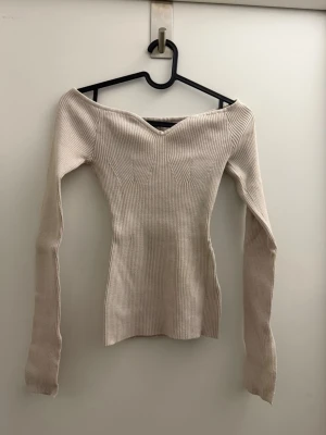 Beige off shoulder stickad tröja - Trendig beige stickad tröja med off shoulder-design och smal passform. Tröjan har långa ärmar och ribbad struktur som ger en snygg siluett. Perfekt för dig som gillar stilrena och moderna plagg.