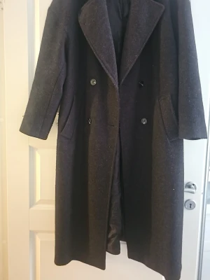 Mörkgrå ullkappa med bred krage - Stilren mörkgrå ullkappa med dubbelknäppning och bred krage. Kappan har två snedställda fickor och är helfodrad för extra komfort. Klassisk rak passform som passar perfekt till höst och vinter.