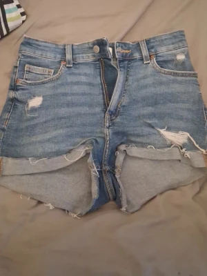 Blå urban jeansshorts med slitningar - Säljer ett par blå jeansshorts med råa kanter och slitna detaljer framt sop locations och bak. Klassisk femficksmodell med knapp och drag coed. Perfekta för en avslappnad och trendig stil i sommar.