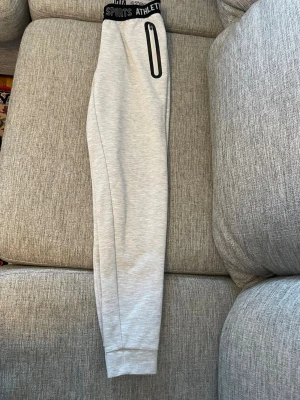 Grå mjukisbyxor från Crivit - Snygga ljusgrå mjukisbyxor från Crivit med bred resår i midjan där det står 'SPORTS ATHLETE'. Byxorna har en rak passform och mudd vid bensluten. Perfekta för chill eller träning, med en enkel och stilren look. Aldrig använd 
