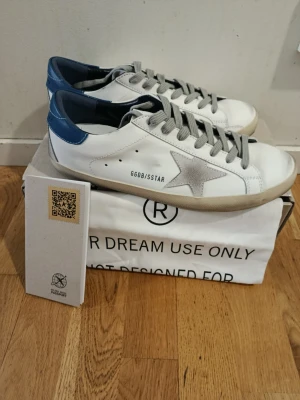Golden Goose vita sneakers med grå stjärna - Golden Goose sneakers i vitt läder med grå mockadetaljer och ikonisk stjärna på sidan. Skorna har grå skosnören, svart hälparti och GGDB/SSTAR-tryck. Klassisk låg modell med slitna detaljer för en cool och avslappnad look.