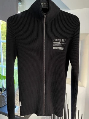 Svart ribbad ziptröja Jack & Jones - Snygg svart ribbad ziptröja från Jack & Jones Core Lab med hög krage och tryck på bröstet. Tröjan har hel dragkedja framtill och är tillverkad i ett mjukt, stretchigt material. Perfekt för en avslappnad och trendig look.