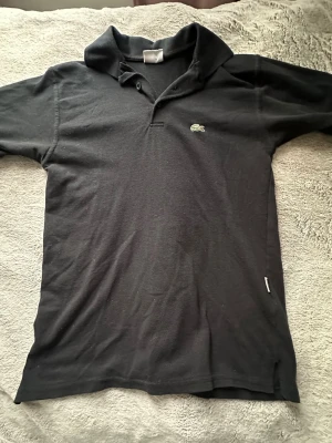 Svart vintidge pikétröja från Lacoste - Klassisk svart pikétröja från Lacoste med korta armar i storlek S.