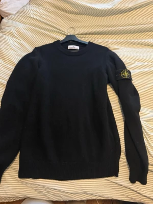 Svart stickad tröja Stone Island - Säljer en svart stickad tröja från Stone Island med den ikoniska logotypen på vänster ärm. Tröjan har rund halsringning, långa ärmar och ribbade muddar. Perfekt för dig som gillar streetwear och vill ha något stilrent och klassiskt.