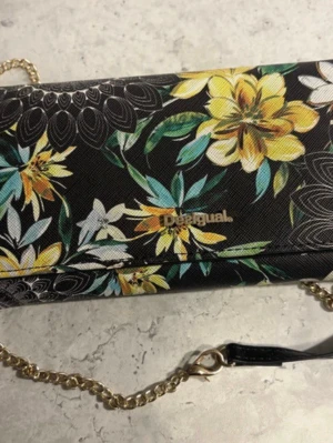 Blommig plånboksväska  från Desigual - Snygg plånboksväska  från Desigual med färgglatt blommönster på svart bakgrund. Väskan har guldfärgad kedjerem och logga framtill. Perfekt för dig som vill sticka ut med en unik accessoar. Materialet är konstläder.