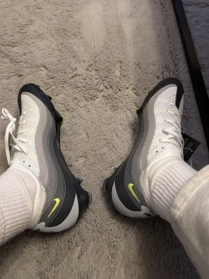 Nike Air fotbollsskor vit/grå - Nike Air fotbollsskor med snygg design i vitt och grått, detaljer i neongult och svart. Skon har snörning, stickad ovandel och inslag av syntet och skinn. Platt sula med dobbar för gräsplan. Perfekt för dig som vill ha stil på planen.