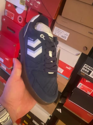 Marinblå Hummel sneakers suede - Snygga marinblå sneakers från Hummel i mocka med klassiska vita pilar på sidan och mörkbruna gummisulor. Skorna har låga skaft och mörkblå snören. Perfekta för dig som gillar retrovibbar och sportig stil.