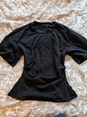 Svart topp med slits i ryggen - En svart topp i bomull med korta ärmar och rund hals. Toppen har en unik detalj med slits baktill som ger en cool twist till den klassiska t-shirten. Passformen är något figurnära och materialet känns mjukt mot huden.