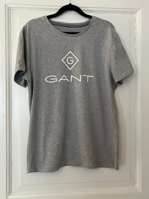 Grå GANT t-shirt med logga - Kommer från fräscht djur och rökfritt hem!    Snygg grå t-shirt från GANT med vit logga tryckt framtill. Klassisk rund halsringning och korta ärmar. Tillverkad i mjuk bomull för en skön känsla och enkel stil som funkar till allt.