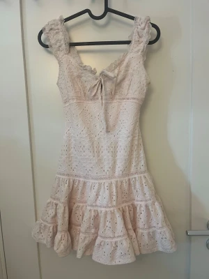 Ljusrosa spetsklänning med volang - Supersöt ljusrosa klänning med breda axelband, volangkant och omlottknytning fram. Den är i storlek XXS 