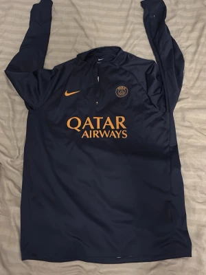Mörkblå PSG Nike Storm-FIT tröja - Snygg mörkblå långärmad träningströja från Nike med PSG-logga och orange detaljer. Tröjan har halv dragkedja, Storm-FIT material och Qatar Airways tryck på bröstet. Perfekt för fotbollsträning eller som supporterplagg.