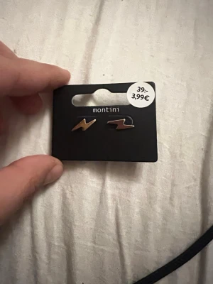 Guldiga blixtörhängen från Montini - Säljer ett par stilrena örhängen från Montini i form av blixtar. De är guldiga och har en cool, minimalistisk design som verkligen sticker ut. Perfekta för dig som vill addera lite edge till din look. Materialet är metall.