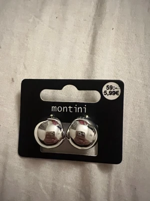Silverfärgade klotörhängen Montini - Säljer ett par stilrena örhängen från Montini i silverfärgad metall. Örhängena har en rund, klotformad design som ger en modern och minimalistisk look. Perfekta för dig som gillar enkla men snygga accessoarer.