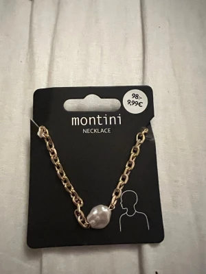Guldfärgat halsband med pärldetalj Montini - Snyggt halsband från Montini med grov guldfärgad kedja och en stor oregelbunden vit pärldetalj i mitten. Perfekt accessoar för att lyfta din outfit med en trendig touch. Kedjan har en modern chunky stil och pärlan ger en cool kontrast. 