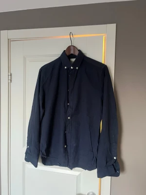 Mörkblå skjorta med knappar från Dobber - Stilren mörkblå skjorta från Dobber med klassisk krage och vita knappar. Skjortan har lång ärm och är tillverkad i ett mjukt bomullstyg som passar perfekt till många olika looks. Enkel och tidlös design som är lätt att matcha. 