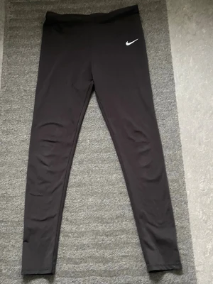 Svarta leggings från Nike - Snygga svarta leggings från Nike med klassisk vit logga på höften. De har en tight passform och är tillverkade i ett stretchigt material som sitter skönt på kroppen. Perfekta för träning eller chill dagar. Midjehöga och utan synliga fickor.
