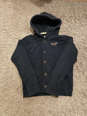 Mörkblå  hoodie från Hollister med knappar - Säljer denna snygga Hollister cardiganen i storlek M. Den har några defekter som man ser på bilderna
