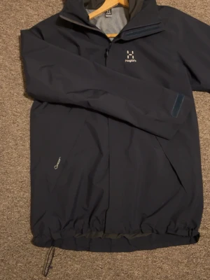 Mörkblå vindjacka från Haglöfs - Snygg mörkblå vindjacka från Haglöfs. Använd ett fåtal gånger ny utan prislapp. med dragkedja framtill och justerbar nederkant. Jackan har hög krage, diskret logga på bröstet och är tillverkad i ett lätt, vindtätt material. Perfekt för blåsiga dagar och outdoor-aktiviteter.