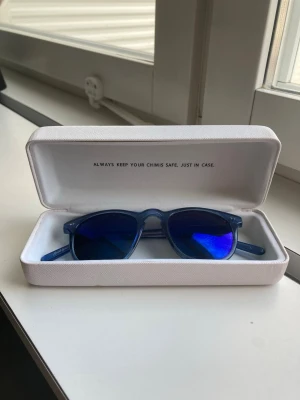 Blåa solglasögon från Chimi - Snygga blåa solglasögon från Chimi med spegelblåa linser och klassisk rundad form. Bågarna är i blått syntetmaterial och kommer med ett stilrent vitt fodral. Perfekt för dig som vill sticka ut med coola accessoarer. Det är en repa på ena glaset som syns på bild