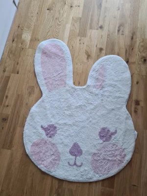 Söt kaninmatta i pastell - Söt matta i form av ett kaninansikte från Kotikulta. Mattan är vit med ljusrosa detaljer på öron, kinder och nos. Tillverkad i mjukt material, sopass stor att den passar perfekt i barnrummet eller som mysig detalj i hemmet.